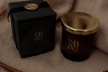 Load image into Gallery viewer, A dark amber glass candle jar with a polished gold lid sits beside its black gift box, both featuring a gold 'WB &' monogram logo. The box is tied with a black ribbon and topped with a gold seal, set against a soft beige fabric background.